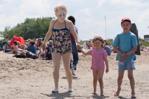 Maskenspielerin mit Badehose geht am Strand spazieren mit Kinder